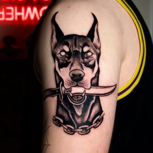 Nahaufnahme eines schwarzen Oberarm-Tattoos mit Dobermann-Motiv, Dolch, Kette und detaillierten Schattierungen.